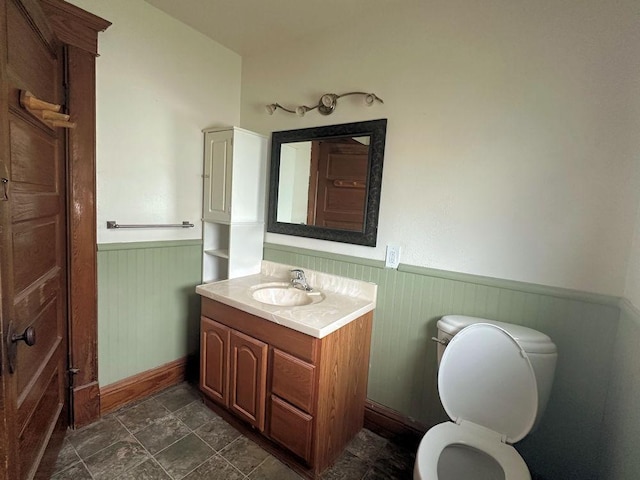bathroom featuring wainscoting, vanity, baseboards, toilet, and stone finish floor