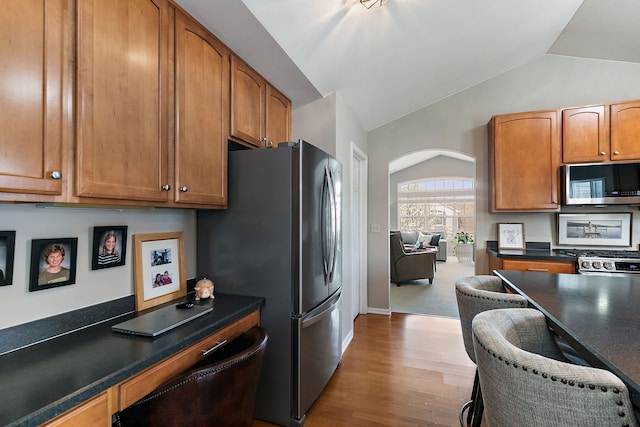 kitchen with dark countertops, appliances with stainless steel finishes, vaulted ceiling, brown cabinets, and wood finished floors