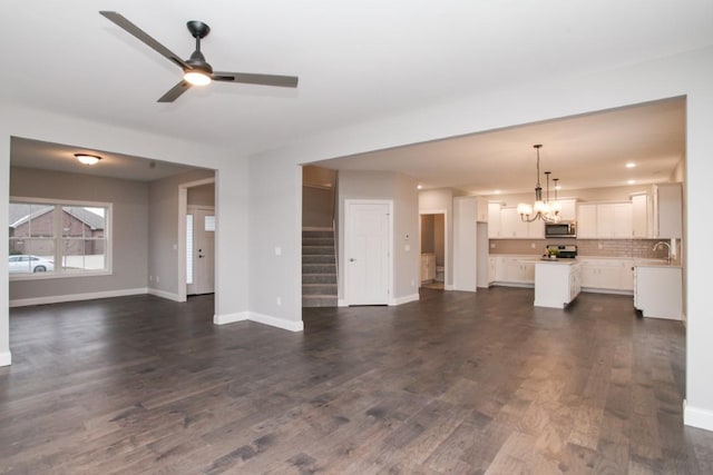 unfurnished living room featuring ceiling fan with notable chandelier, baseboards, stairway, and dark wood finished floors