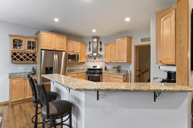 kitchen featuring wall chimney range hood, stainless steel appliances, a kitchen breakfast bar, and light brown cabinetry