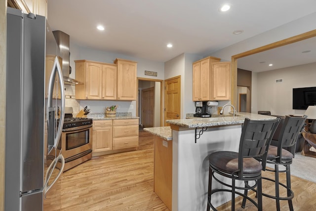 kitchen with light brown cabinets, light wood-style flooring, a breakfast bar area, stainless steel appliances, and wall chimney range hood