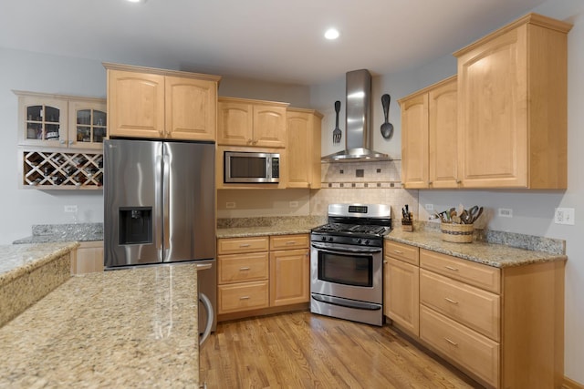 kitchen featuring light stone countertops, light wood-style floors, light brown cabinetry, wall chimney range hood, and appliances with stainless steel finishes