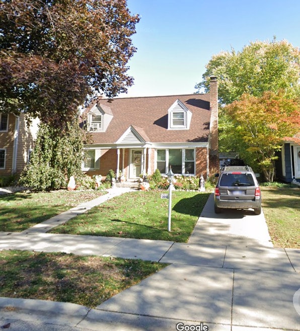 cape cod-style house with concrete driveway, a front yard, a chimney, and brick siding