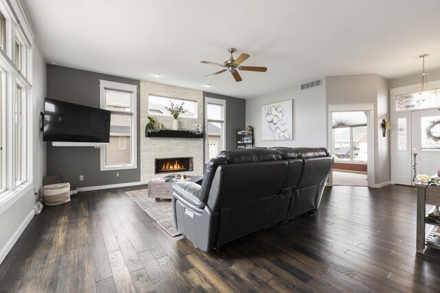 living room featuring a stone fireplace, ceiling fan, visible vents, baseboards, and dark wood-style floors