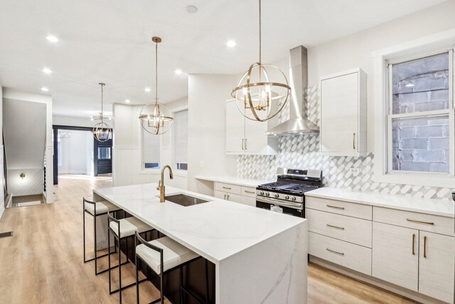 kitchen with a sink, a kitchen island with sink, an inviting chandelier, wall chimney exhaust hood, and stainless steel gas range oven