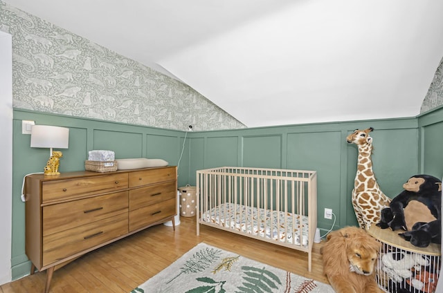 bedroom featuring lofted ceiling, a decorative wall, wallpapered walls, a crib, and wood finished floors