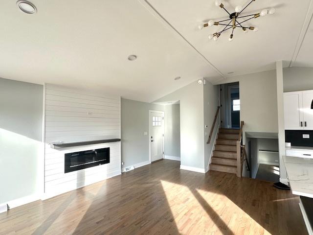 unfurnished living room featuring a glass covered fireplace, vaulted ceiling, wood finished floors, and stairway