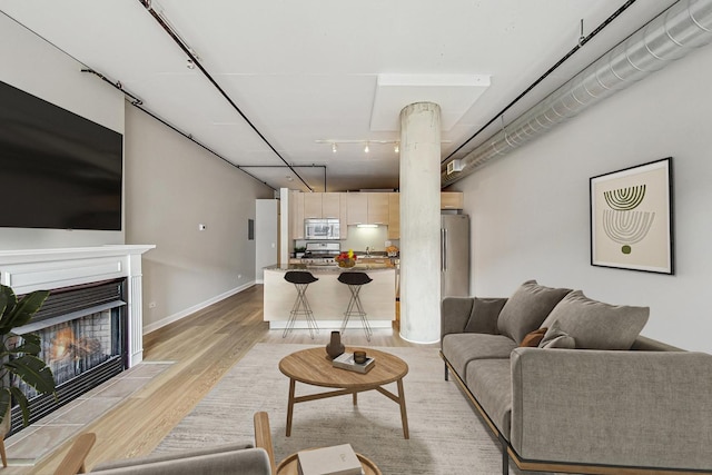 living area featuring light wood-type flooring, decorative columns, baseboards, a warm lit fireplace, and rail lighting