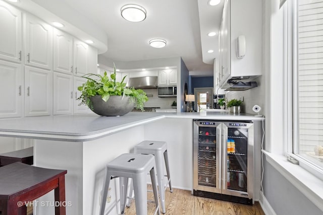 bar featuring wall chimney range hood, stainless steel microwave, light wood finished floors, beverage cooler, and recessed lighting