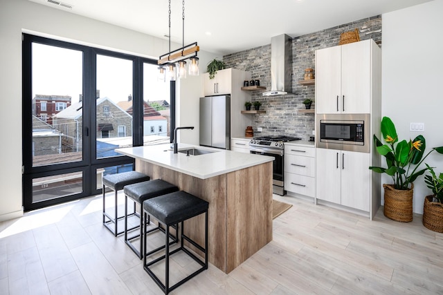kitchen featuring stainless steel appliances, wall chimney range hood, backsplash, a sink, and a center island with sink