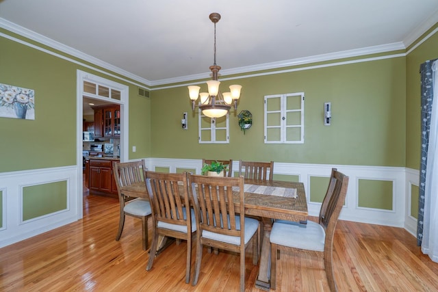 dining space with light wood-style flooring, ornamental molding, a chandelier, wainscoting, and a decorative wall