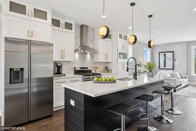 kitchen with dark wood finished floors, a sink, wall chimney range hood, a kitchen island with sink, and appliances with stainless steel finishes