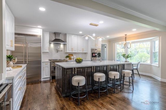 kitchen with wall chimney range hood, high quality appliances, white cabinets, a breakfast bar, and light countertops