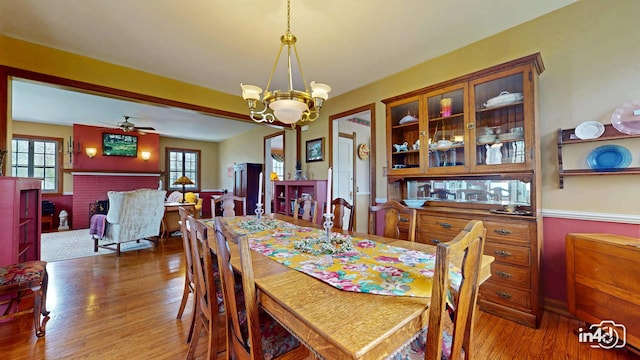 dining space with wood finished floors, ceiling fan with notable chandelier, and a brick fireplace