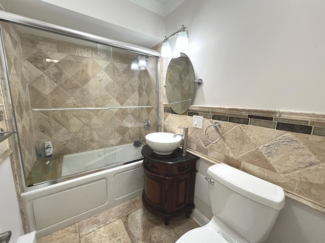 full bath featuring stone tile flooring, toilet, tile walls, combined bath / shower with glass door, and vanity