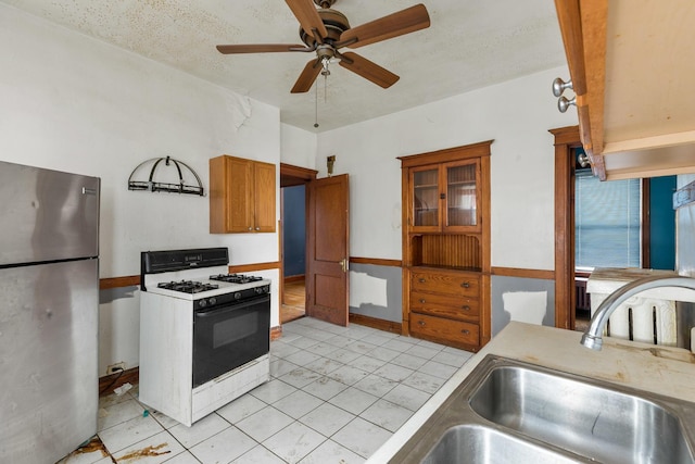 kitchen featuring freestanding refrigerator, brown cabinets, gas range oven, ceiling fan, and a sink