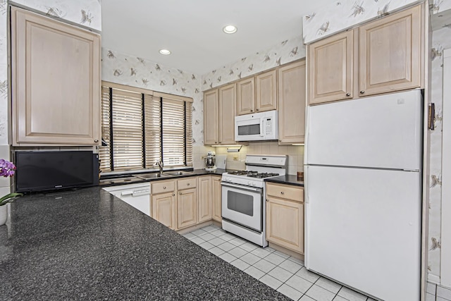 kitchen with wallpapered walls, dark countertops, light brown cabinets, a sink, and white appliances