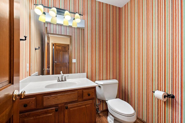 half bathroom featuring wallpapered walls, wood finished floors, toilet, and vanity