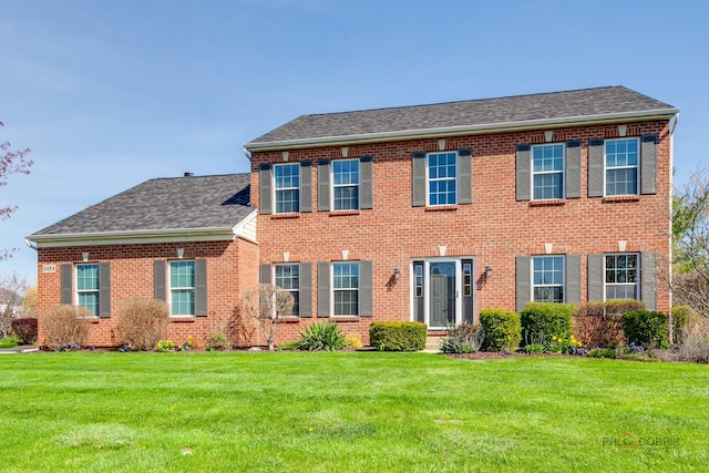 colonial house with brick siding, a front lawn, and roof with shingles