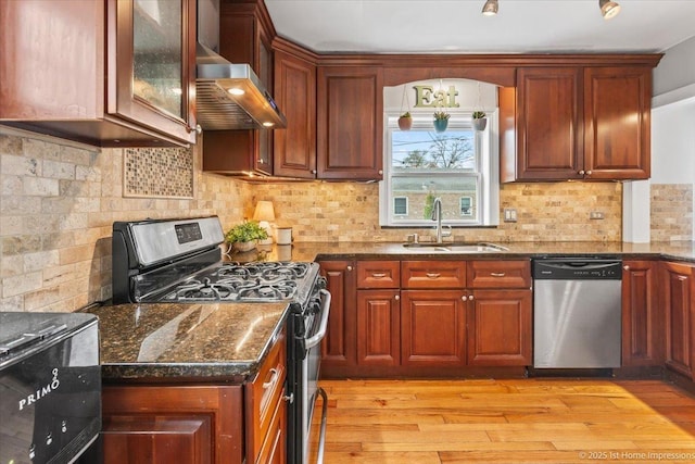 kitchen featuring appliances with stainless steel finishes, a sink, light wood finished floors, dark stone counters, and wall chimney exhaust hood