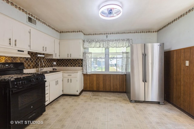 kitchen with black gas range, freestanding refrigerator, wood walls, under cabinet range hood, and a sink