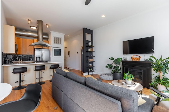 living area with crown molding, light wood finished floors, a glass covered fireplace, recessed lighting, and visible vents