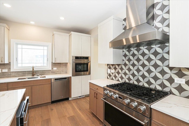 kitchen featuring a sink, decorative backsplash, light wood-style flooring, wall chimney range hood, and appliances with stainless steel finishes