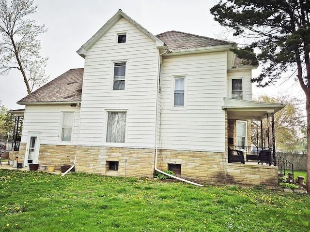 rear view of house featuring a lawn, roof with shingles, covered porch, and stone siding
