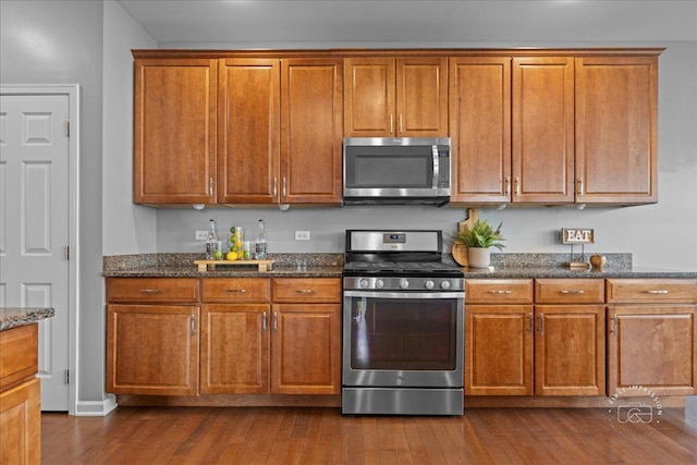 kitchen featuring dark wood-style flooring, stainless steel appliances, brown cabinetry, and dark stone counters