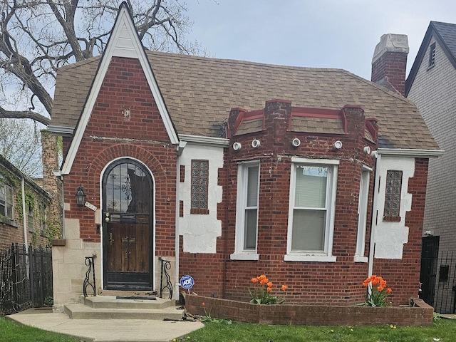 tudor home featuring brick siding, roof with shingles, fence, and a chimney