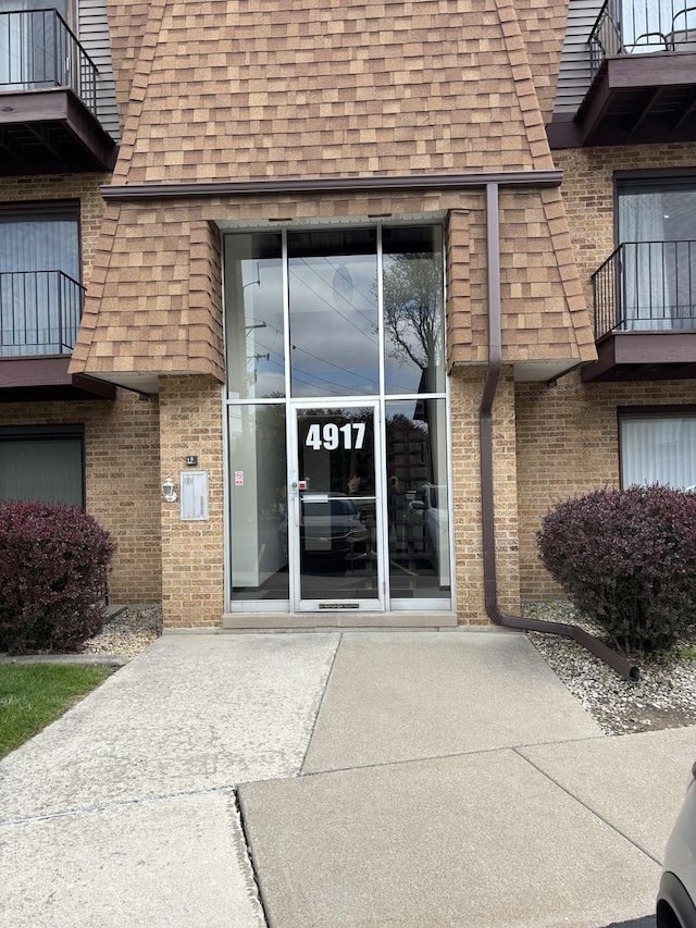 doorway to property featuring a shingled roof, mansard roof, brick siding, and a balcony