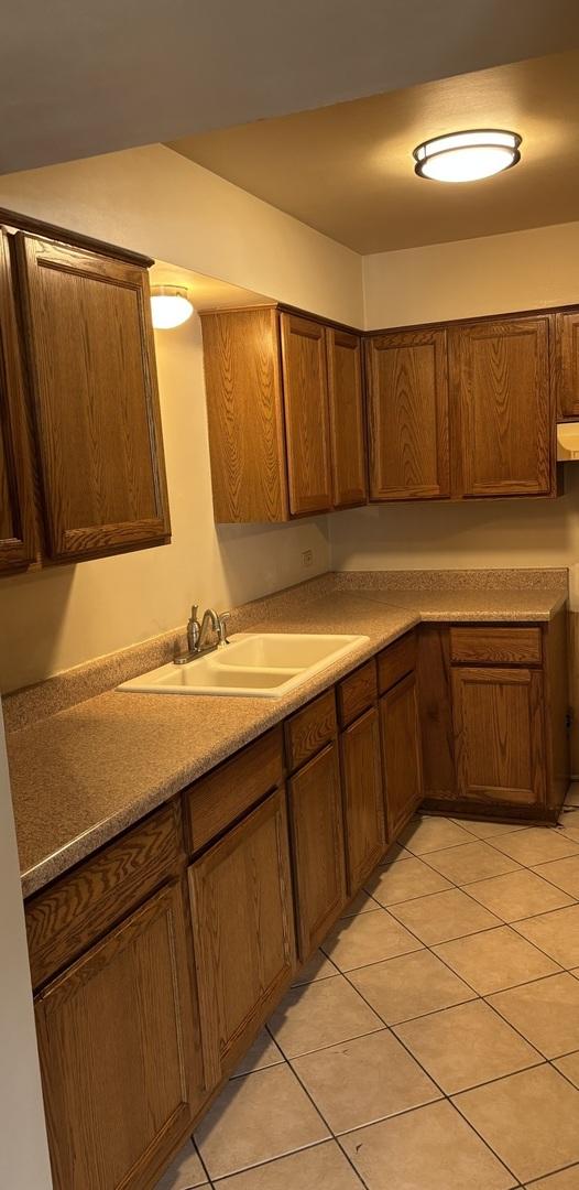 kitchen with light tile patterned floors, a sink, brown cabinetry, under cabinet range hood, and light countertops