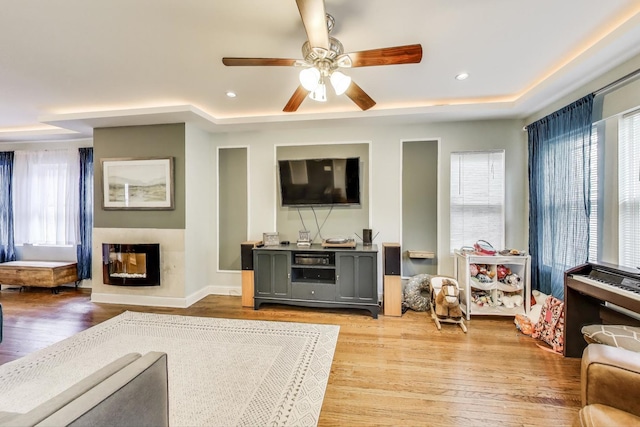 living area with a multi sided fireplace, ceiling fan, a tray ceiling, recessed lighting, and light wood finished floors
