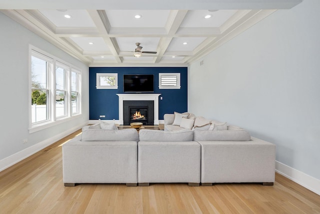 living area featuring beamed ceiling, a glass covered fireplace, a ceiling fan, baseboards, and light wood-style floors