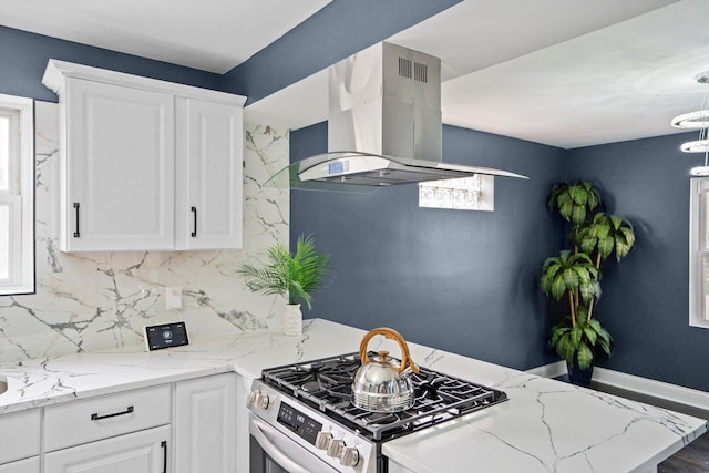 kitchen with light stone countertops, stainless steel gas range oven, wall chimney range hood, white cabinets, and backsplash