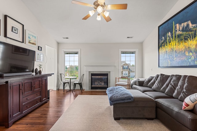 living area featuring dark wood-type flooring, a ceiling fan, a fireplace with flush hearth, and visible vents