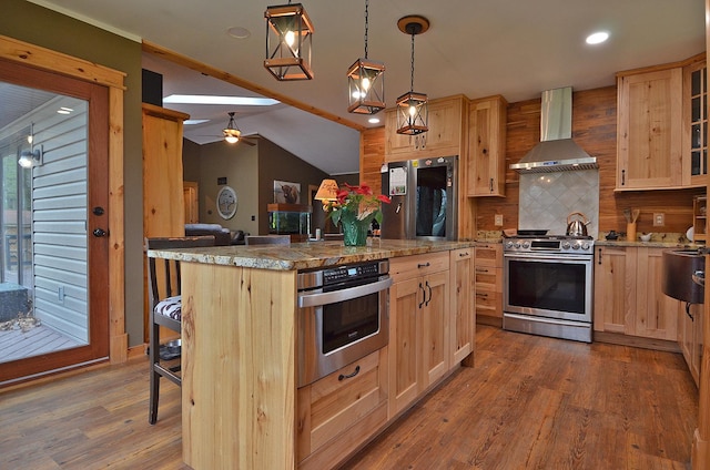 kitchen with light brown cabinetry, wood finished floors, appliances with stainless steel finishes, and wall chimney range hood