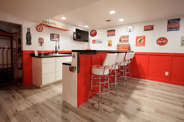 bar with light wood-style floors, a wainscoted wall, indoor bar, and a sink