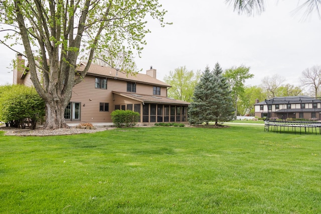 back of property with a sunroom, a trampoline, a chimney, a shingled roof, and a yard