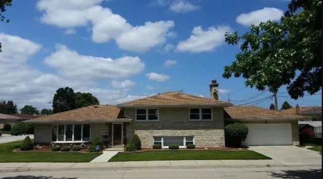 split level home featuring driveway, stone siding, a chimney, a front yard, and a garage