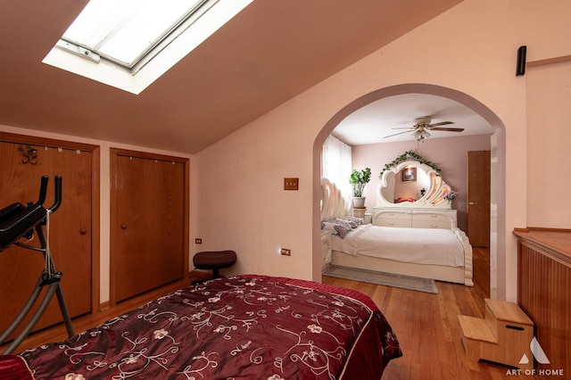 bedroom featuring vaulted ceiling with skylight, ceiling fan, two closets, arched walkways, and wood finished floors