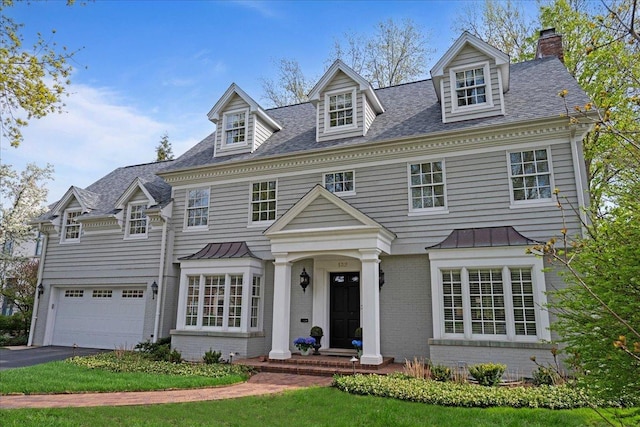 colonial home featuring aphalt driveway, a chimney, a standing seam roof, brick siding, and an attached garage