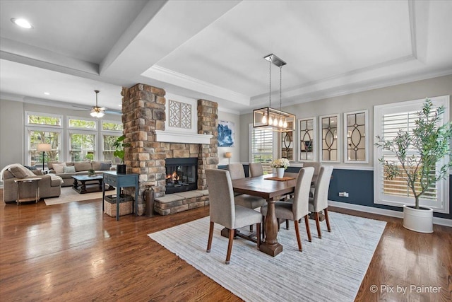 dining area with a raised ceiling, a fireplace, ceiling fan with notable chandelier, and dark wood-style flooring