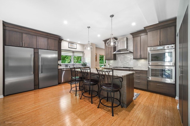 kitchen with wall chimney range hood, dark brown cabinets, a kitchen breakfast bar, stainless steel appliances, and tasteful backsplash