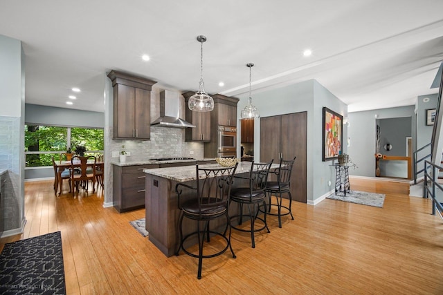 kitchen featuring dark brown cabinetry, a breakfast bar, light wood-style flooring, wall chimney exhaust hood, and baseboards