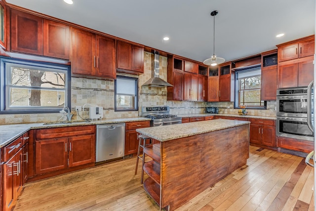 kitchen featuring reddish brown cabinets, light wood-style flooring, appliances with stainless steel finishes, wall chimney range hood, and a sink