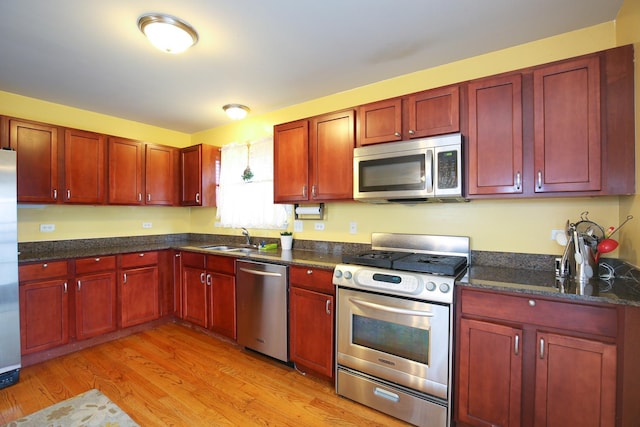 kitchen featuring reddish brown cabinets, dark stone counters, stainless steel appliances, a sink, and light wood-type flooring