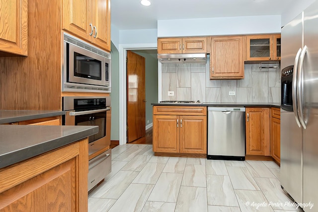 kitchen featuring dark countertops, tasteful backsplash, brown cabinets, glass insert cabinets, and stainless steel appliances