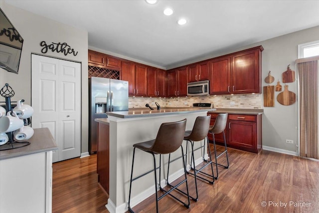 kitchen with wood finished floors, a breakfast bar, reddish brown cabinets, tasteful backsplash, and appliances with stainless steel finishes