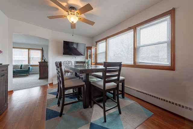 dining space with a ceiling fan, arched walkways, a baseboard heating unit, and wood finished floors